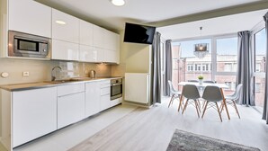 Una cocina moderna con armarios blancos, un microondas y un horno. Hay una zona de comedor con una mesa y sillas, y un ventanal con cortinas.