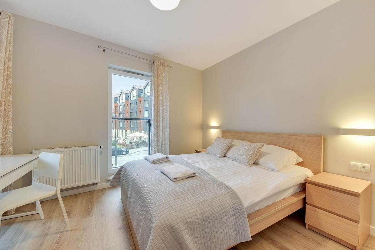 Une chambre avec un grand lit, un bureau et une chaise, une fenêtre avec des rideaux et une vue sur les bâtiments environnants.
