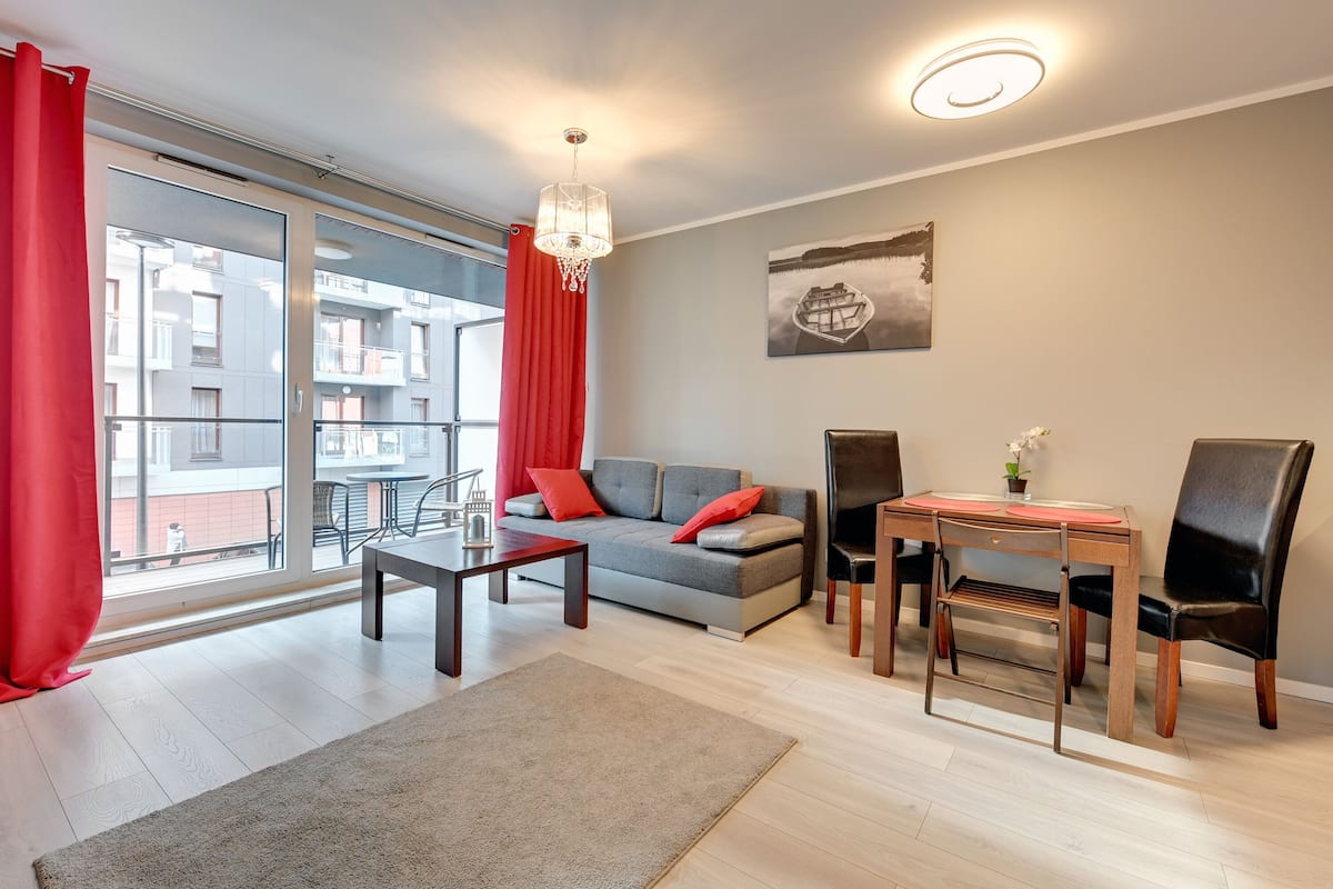 Un salon moderne avec un canapé gris, des coussins rouges, une table basse en bois et une table à manger avec des chaises.