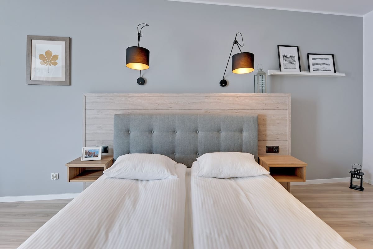 Un lit double avec une tête de lit rembourrée de couleur grise, deux tables de chevet et deux lampes fixées au mur.