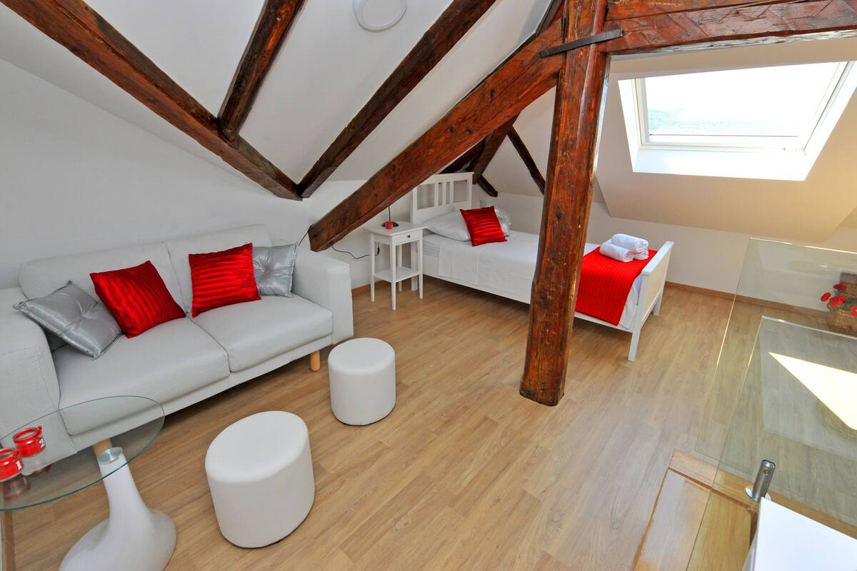 Ein modernes Schlafzimmer im Loftsstil mit schrägverdeckter Decke, einem weißen Bett, einem grauen Sofa mit roten und silbernen Kissen, einem kleinen weißen Tisch und einem Glastisch.