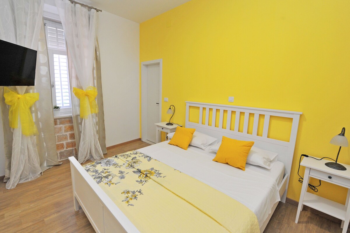 A bedroom with a bed, a nightstand, a lamp, a TV, and a window with curtains.