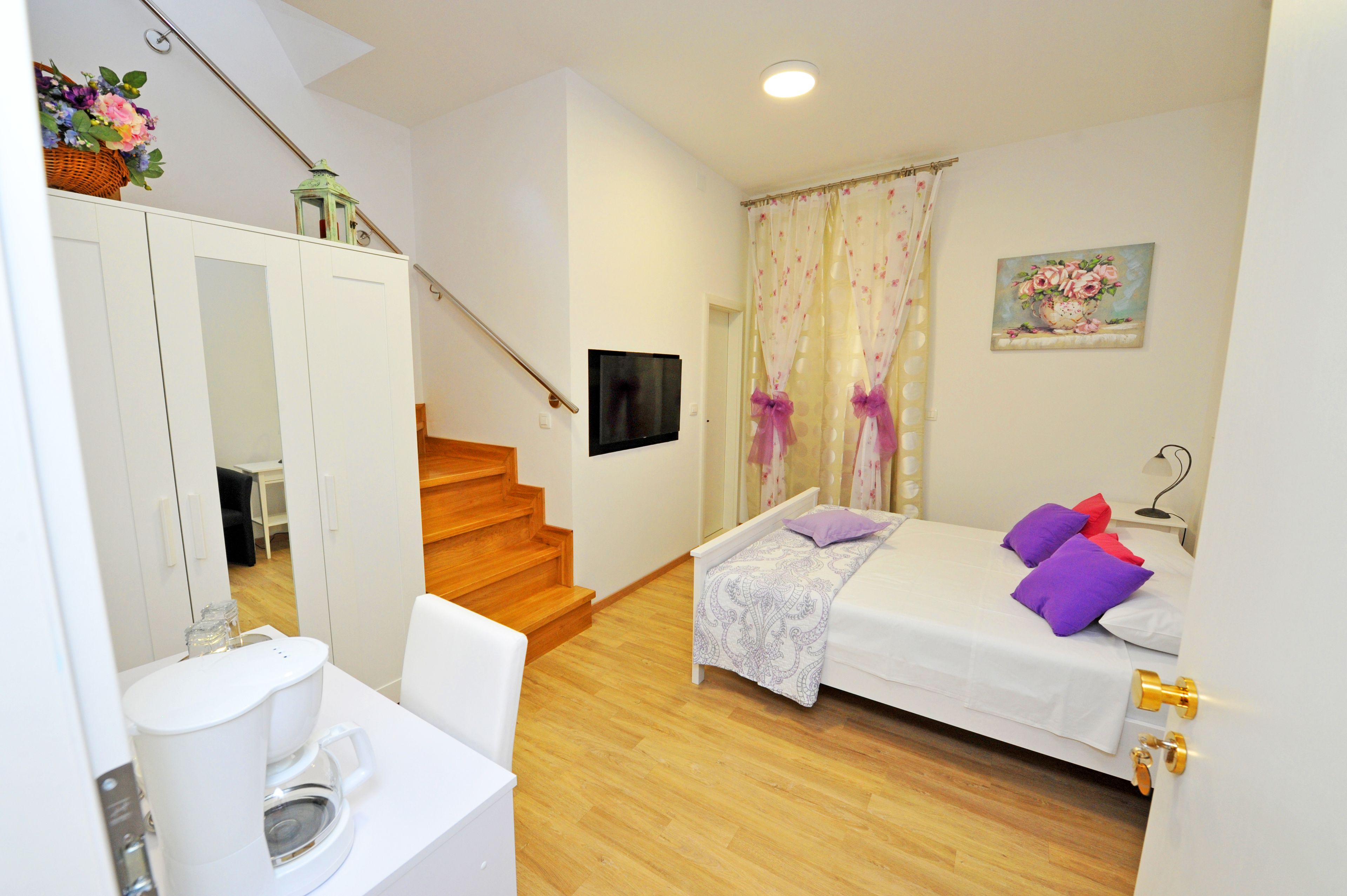 A bedroom with a bed, a TV, a window with curtains, a staircase, and a bathroom with a toilet and sink.