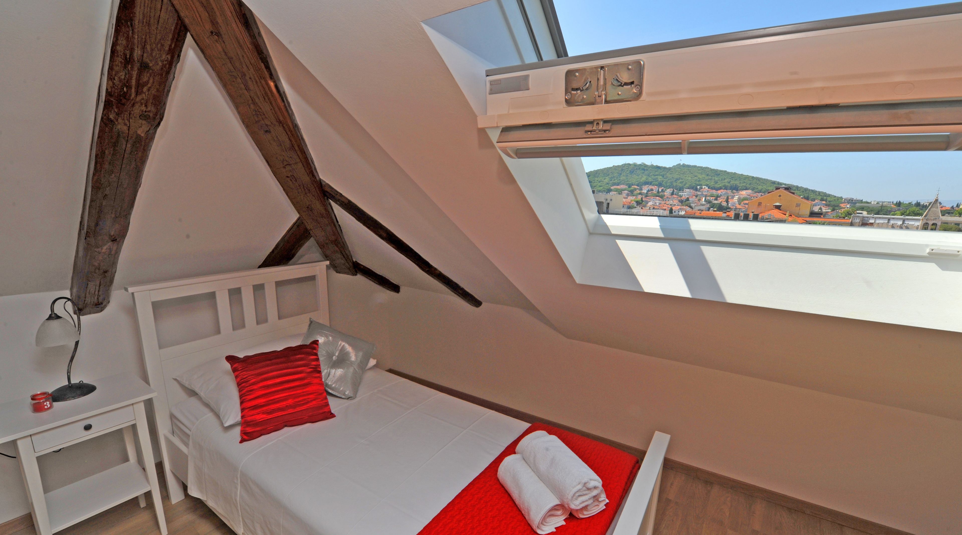 Quarto com teto inclinado, cama com travesseiro vermelho, mesa de cabeceira com abajur e vista de uma cidade por um claraboia.