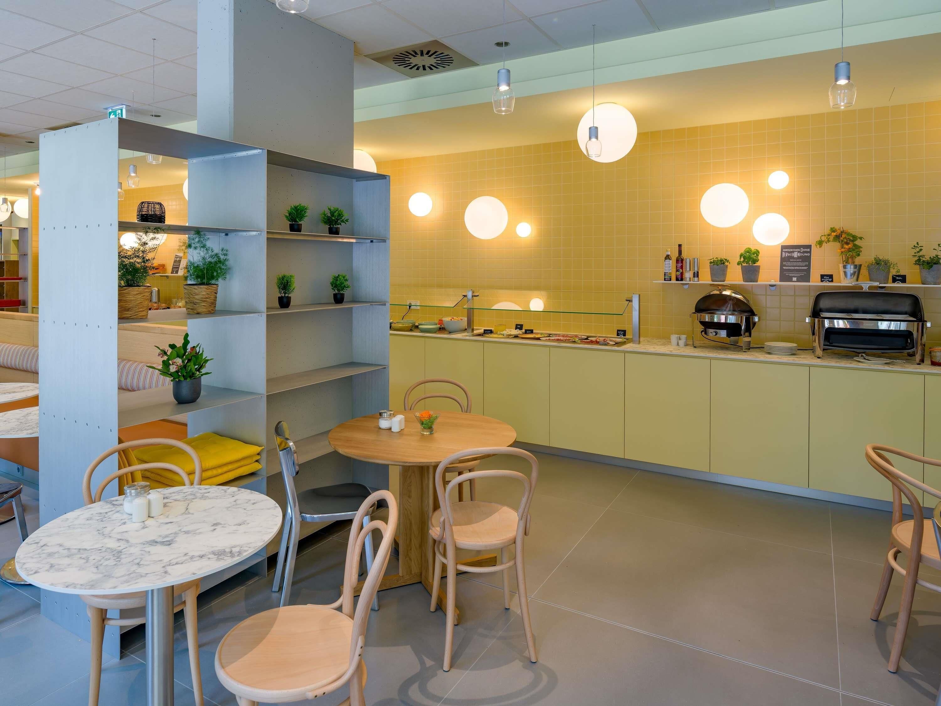 Una cafetería moderna con mesas redondas, sillas de madera, una barra con parrilla y estantes con plantas.