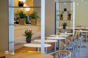 A restaurant interior with round tables, chairs, a wooden bench, and potted plants. - ibis Leipzig City (Leipzig)