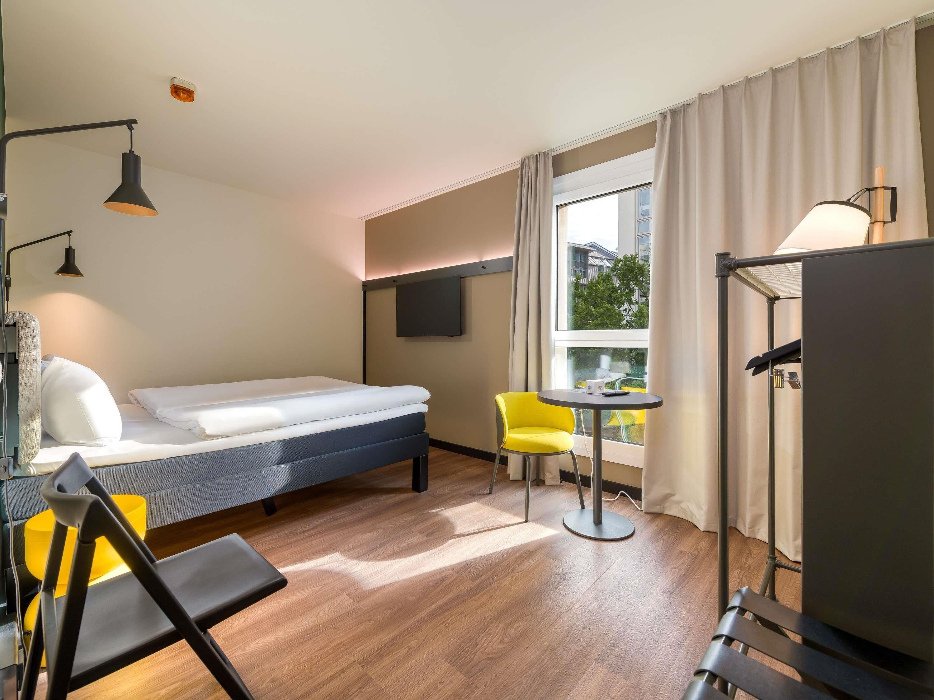 Una camera d'albergo con un letto, un piccolo tavolo, una sedia, una TV e una finestra con tende.