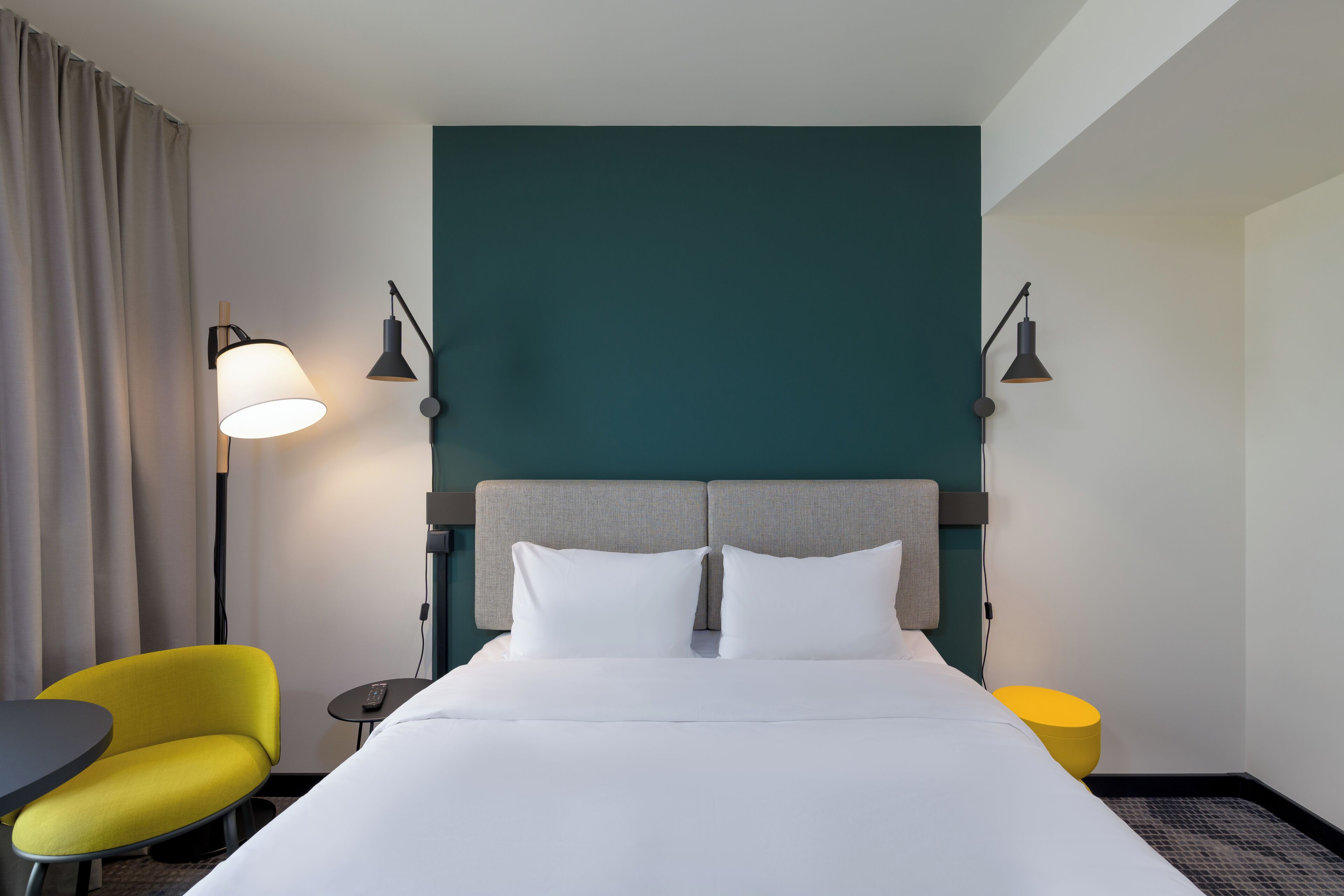 Una habitación de hotel con una cama, dos lámparas de noche, una silla, una mesita y un taburete amarillo.