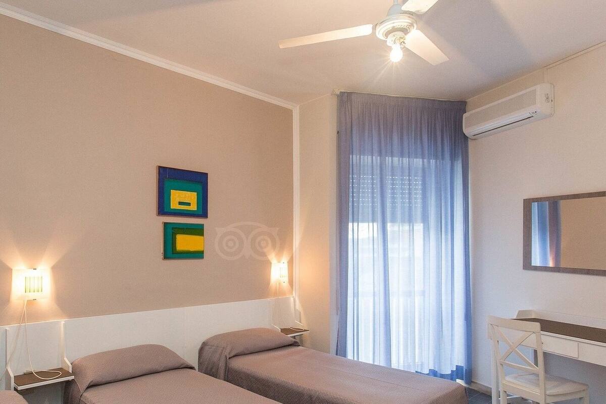 Ein Hotelzimmer mit zwei Betten, einem Schreibtisch, einem Deckenventilator und einem Fenster mit Vorhängen.