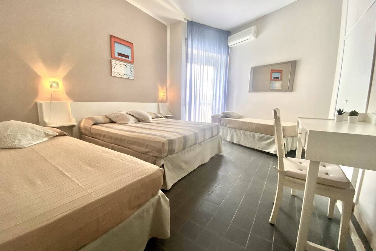 Ein Hotelzimmer mit zwei Betten, einem Schreibtisch und einem Sessel. Es gibt Wandlampen und eine Klimaanlage.