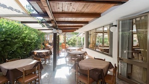 An outdoor patio area with multiple tables and chairs, wooden ceiling beams, and large windows offering views of greenery. - Hotel Anna (Terracina)