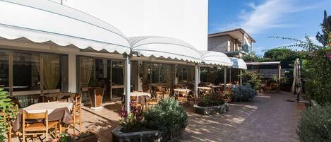 An outdoor patio area with tables and chairs under white umbrellas, surrounded by potted plants and a brick pathway.