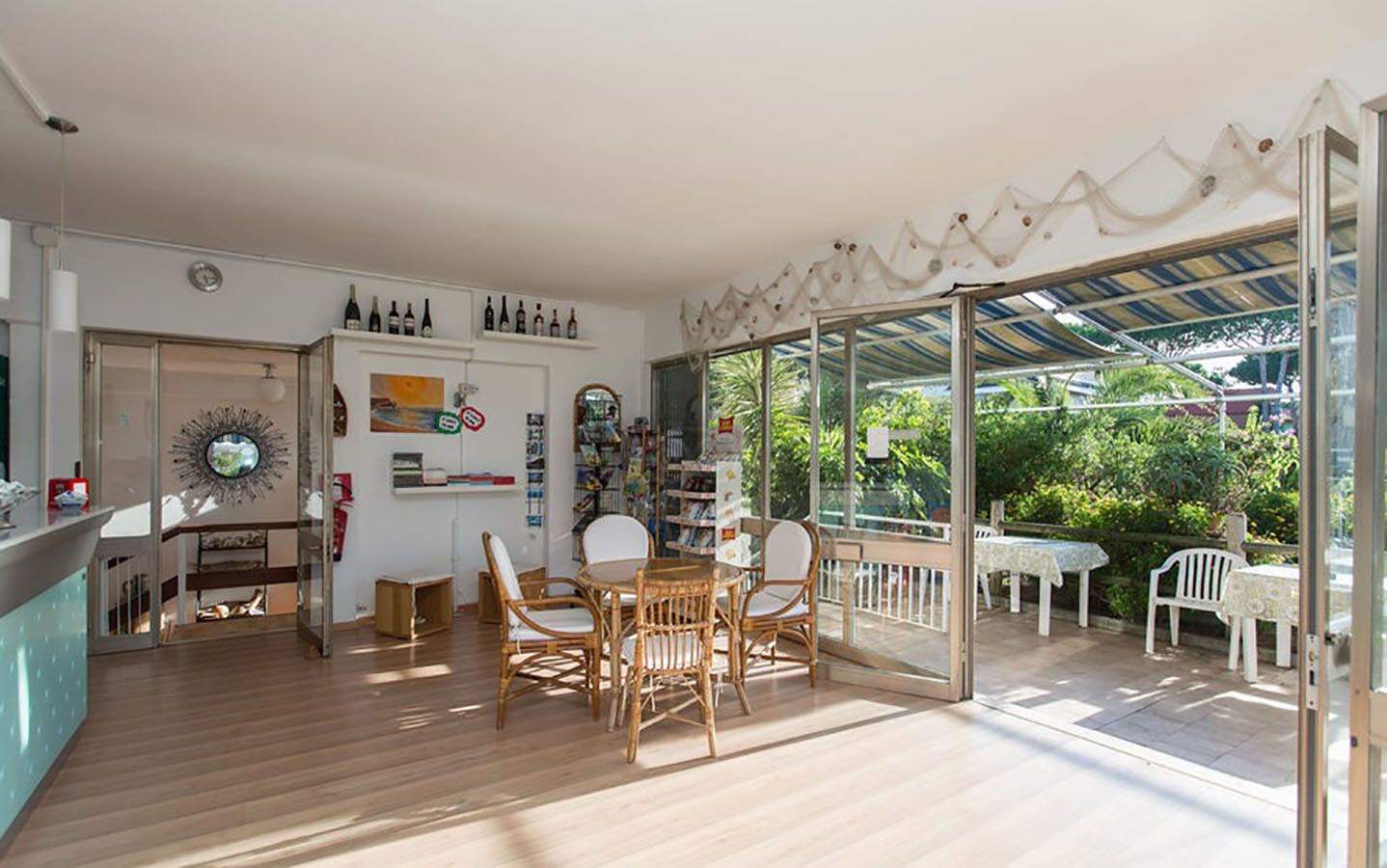 A spacious room with a wooden floor, a dining area with wicker chairs, a bookshelf, and a glass door leading to an outdoor patio with a table and chairs.