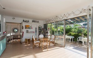 Una habitación amplia con suelo de madera, una zona de comedor con sillas de mimbre, una estantería y una puerta de cristal que da a un patio exterior con mesa y sillas.