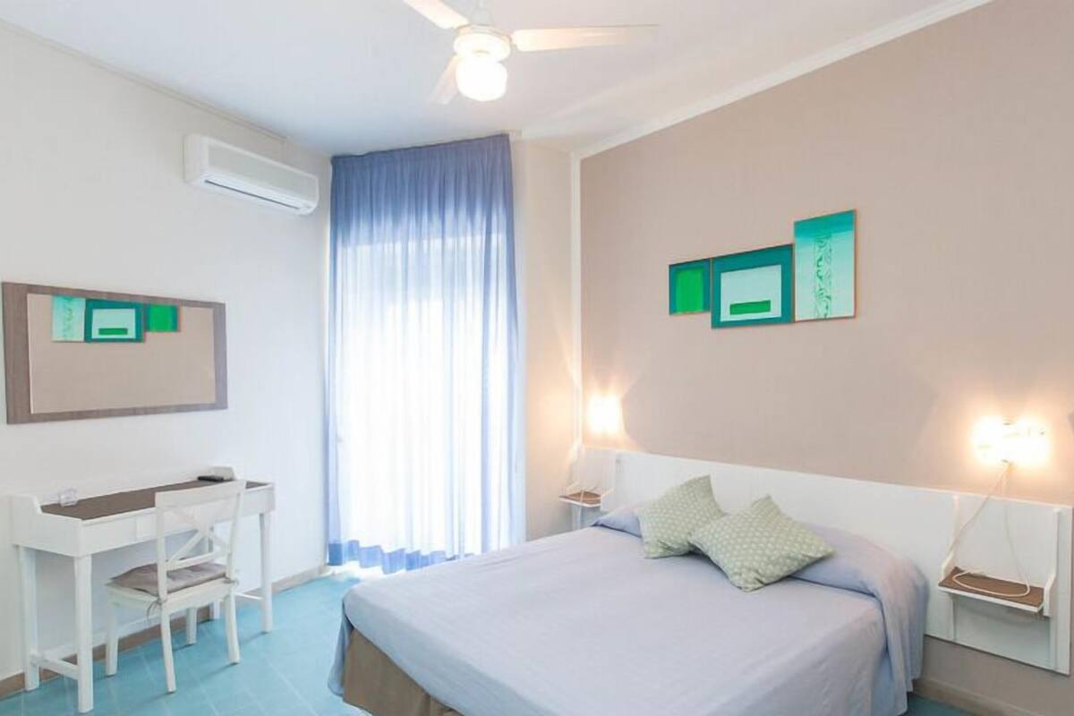 Ein Hotelzimmer mit einem Bett, einem Schreibtisch und einem Stuhl. Es gibt einen Deckenventilator, ein Fenster mit Vorhängen und Wandleuchten.