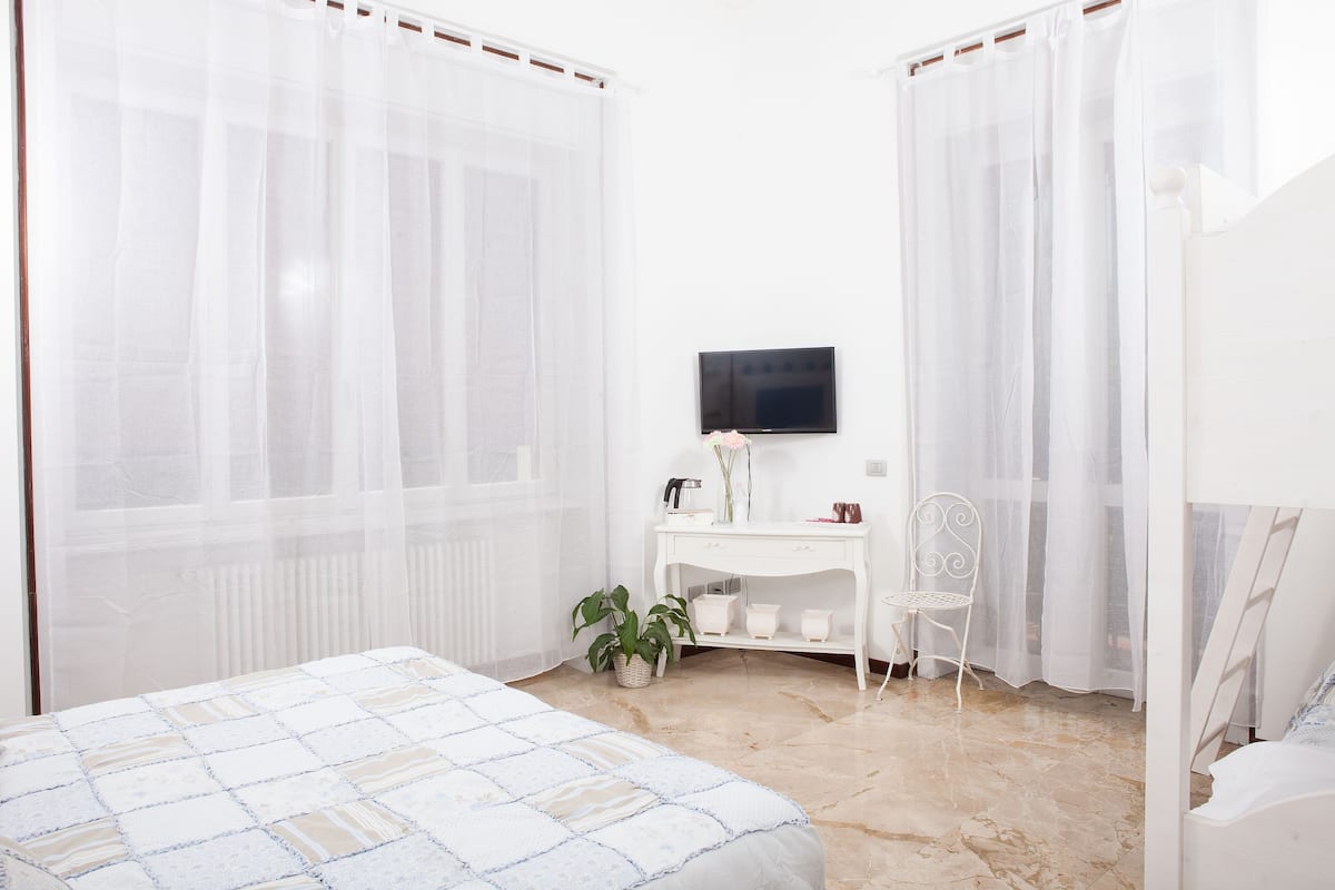 Quarto com cama, escrivaninha, cadeira, televisão e janela com cortinas translúcidas.