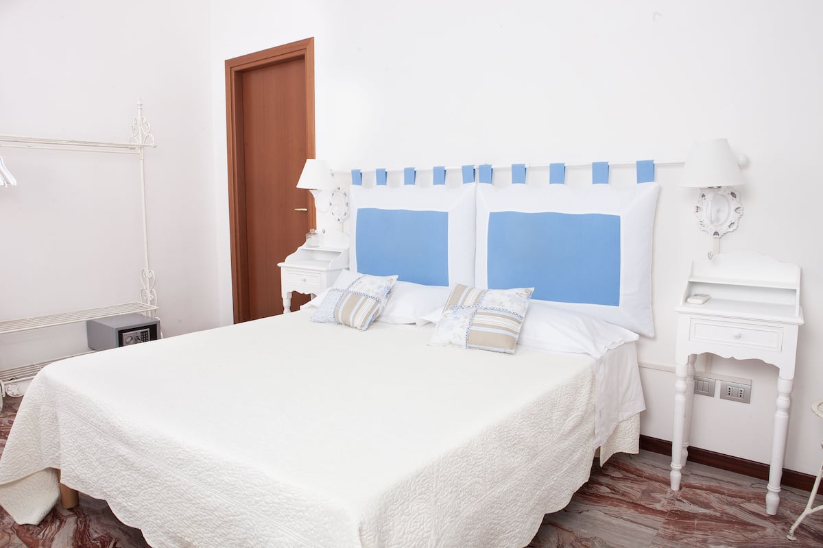 Cama bem arrumada com colcha branca, dois travesseiros com estampas azuis e brancas, mesa de cabeceira com abajur e criado-mudo com relógio.