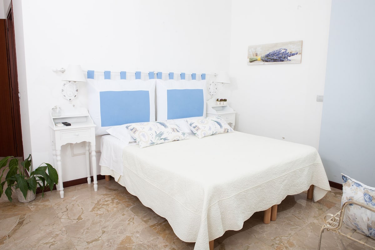 Quarto com cama, mesas de cabeceira, luminária na parede, planta em vaso e uma pintura.