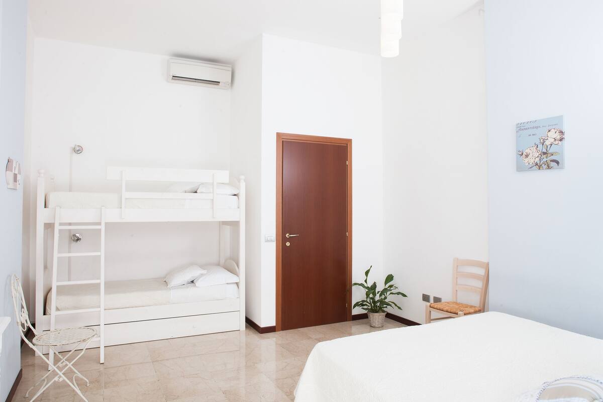 Quarto com cama de solteiro e beliche, porta de madeira, mesa pequena, cadeira e planta em vaso.