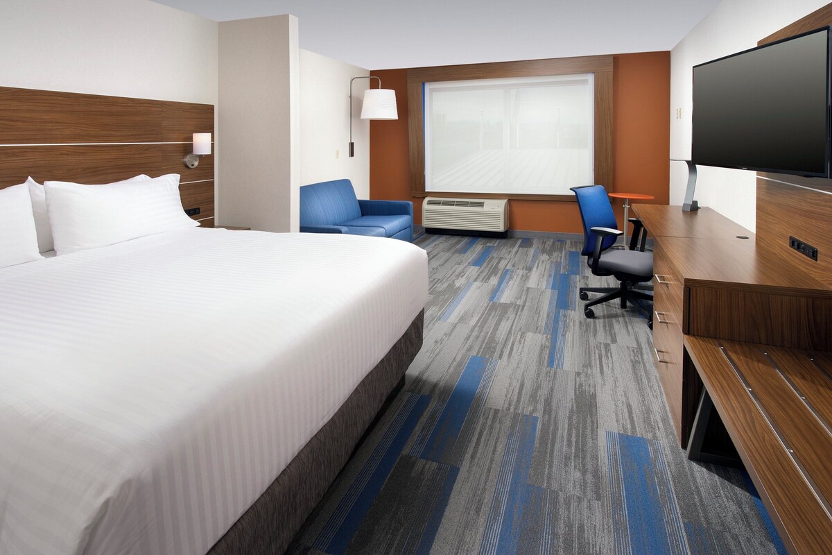 A hotel room with a large bed, a desk with a chair, a flat-screen TV, and a window with blinds.