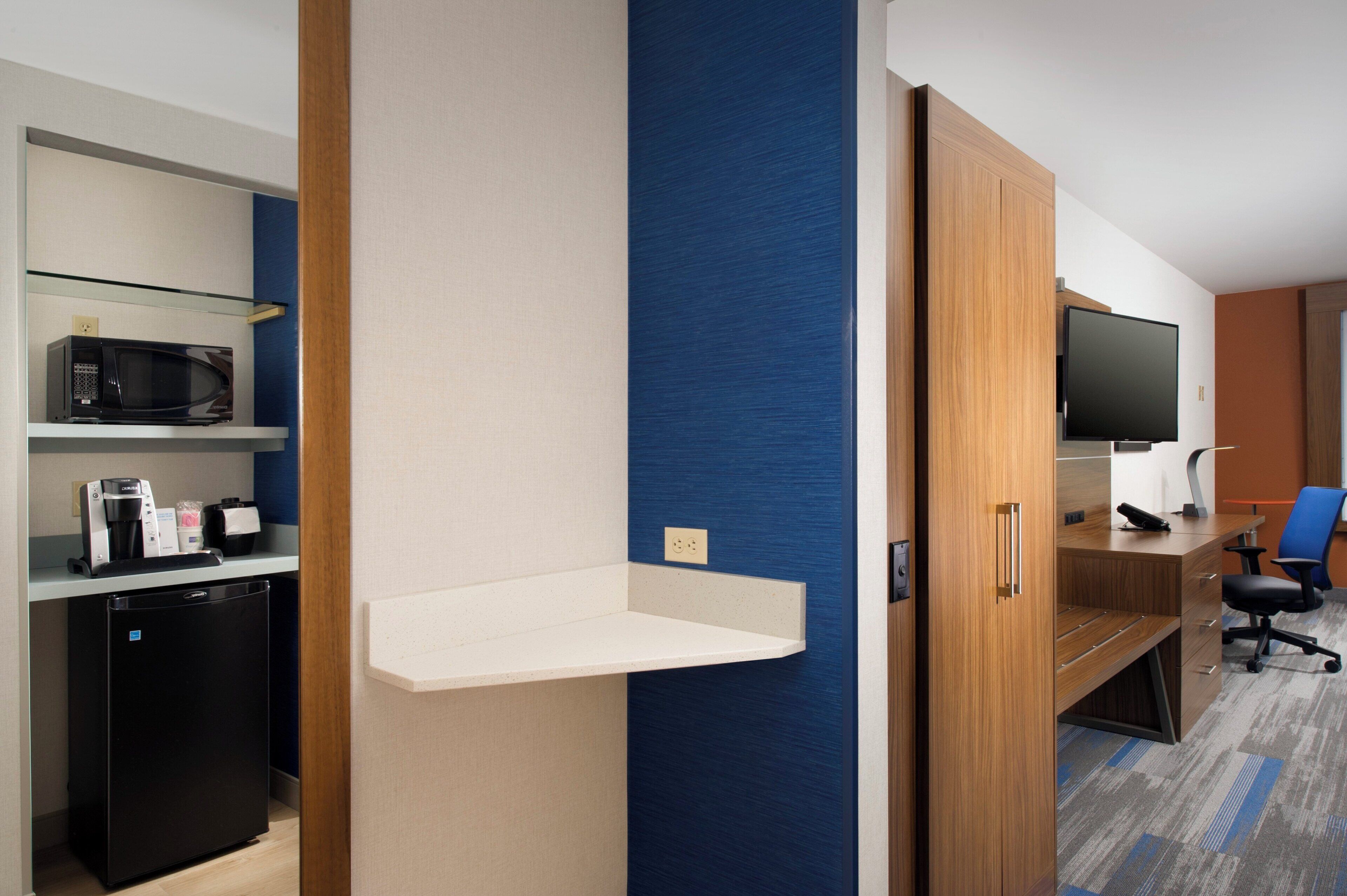 A hotel room with a kitchenette, a flat-screen TV, a desk, and a blue accent wall.