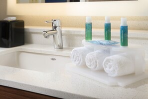 A bathroom sink with a white countertop, a silver faucet, and three bottles of white conditioner and shampoo. - Holiday Inn Express & Suites Altoona by IHG (Altoona)