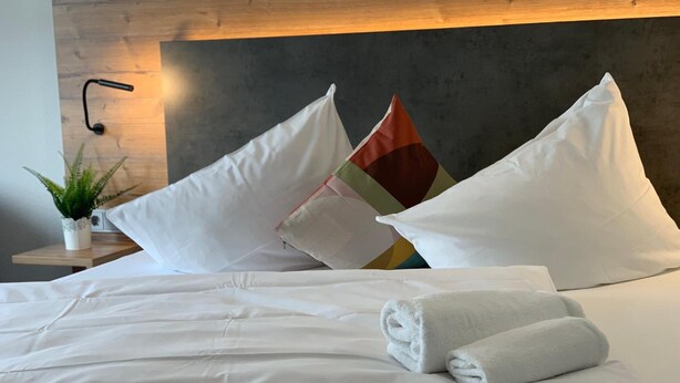 Una cama bien tendida con sábanas blancas, una toalla enrollada, un cojín colorido y una planta de interior.