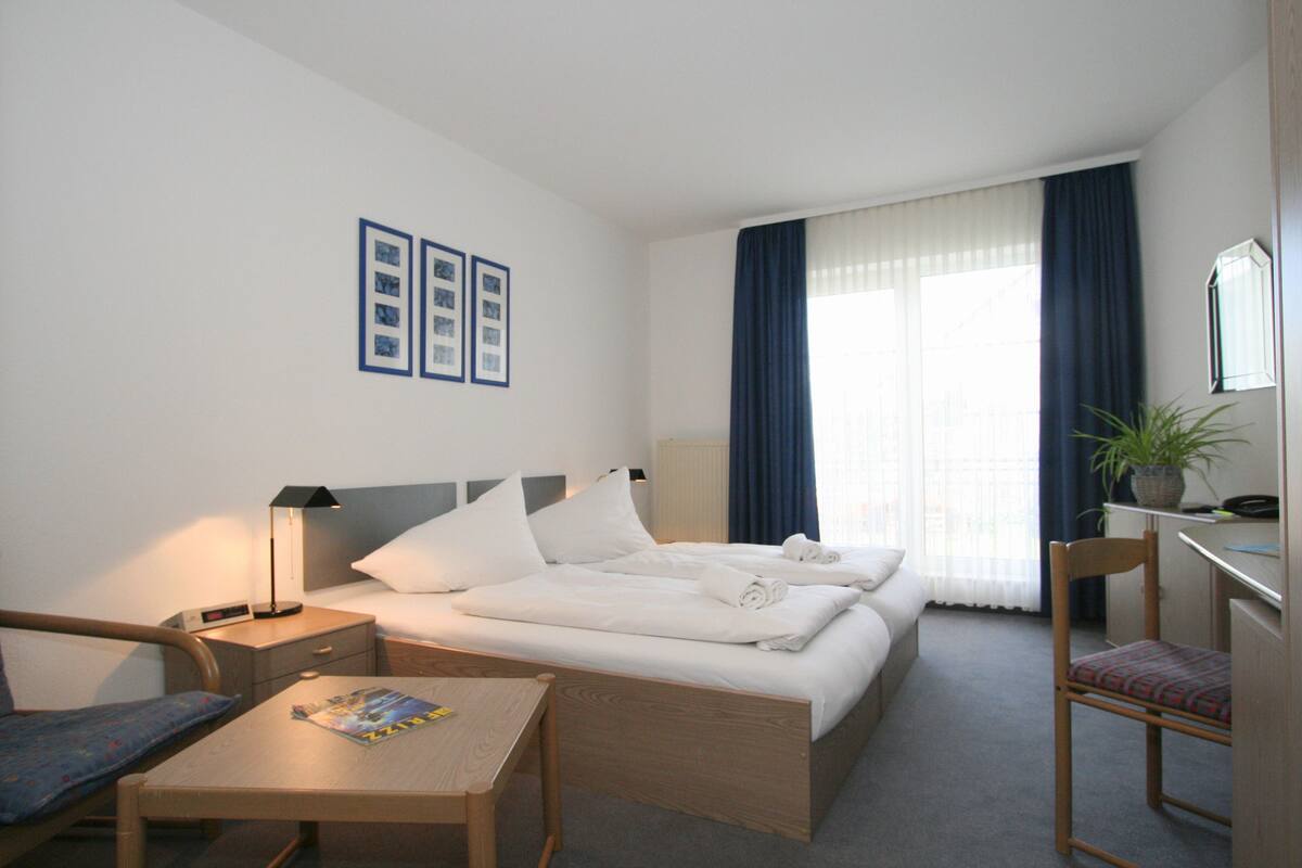 Une chambre d’hôtel avec un lit, deux tables de chevet, une chaise, un bureau et une plante en pot.