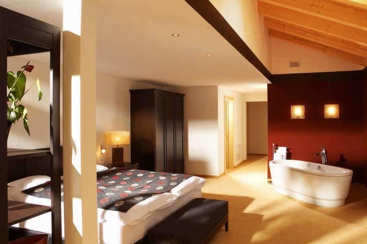 A modern bedroom with a large bed, a dark wardrobe, a bedside lamp, and a bathroom with a bathtub and a red wall.