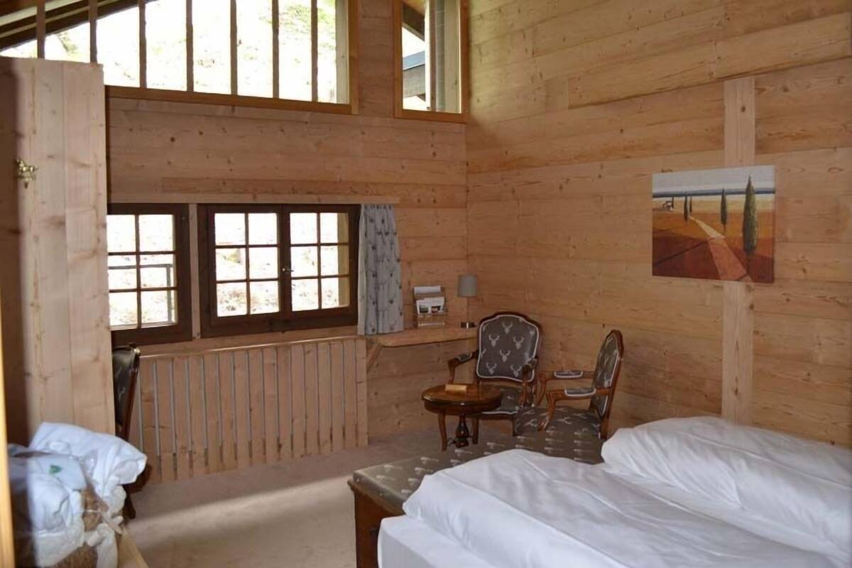 A wooden cabin room with a bed, a chair, a table, a window, and a painting.