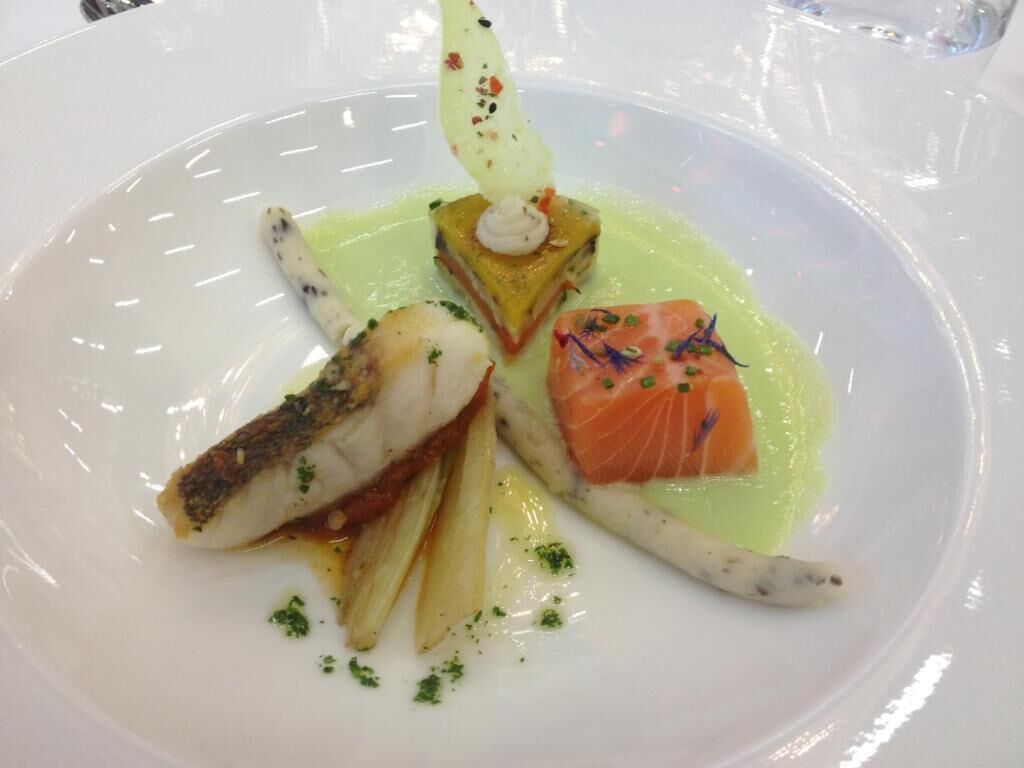 Un plato de comida con varios guisos, incluyendo un trozo de salmón y un pescado blanco, decorado con hierbas.