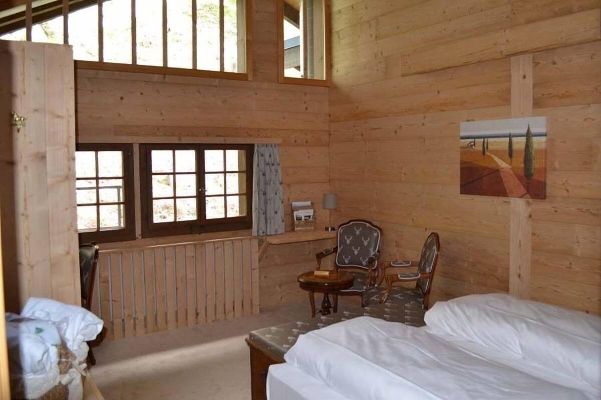 A wooden cabin room with a bed, a chair, a table, and a window.