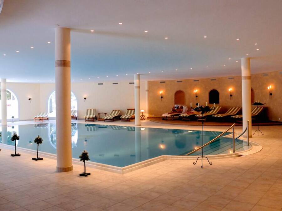 A large indoor swimming pool with lounge chairs and sun loungers around it, surrounded by columns and a tiled floor.