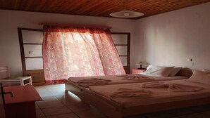 Un dormitori amb sostre de fusta, un llit amb roba de llit blanca, una nitòria, una cadira i una finestra amb cortines florals.