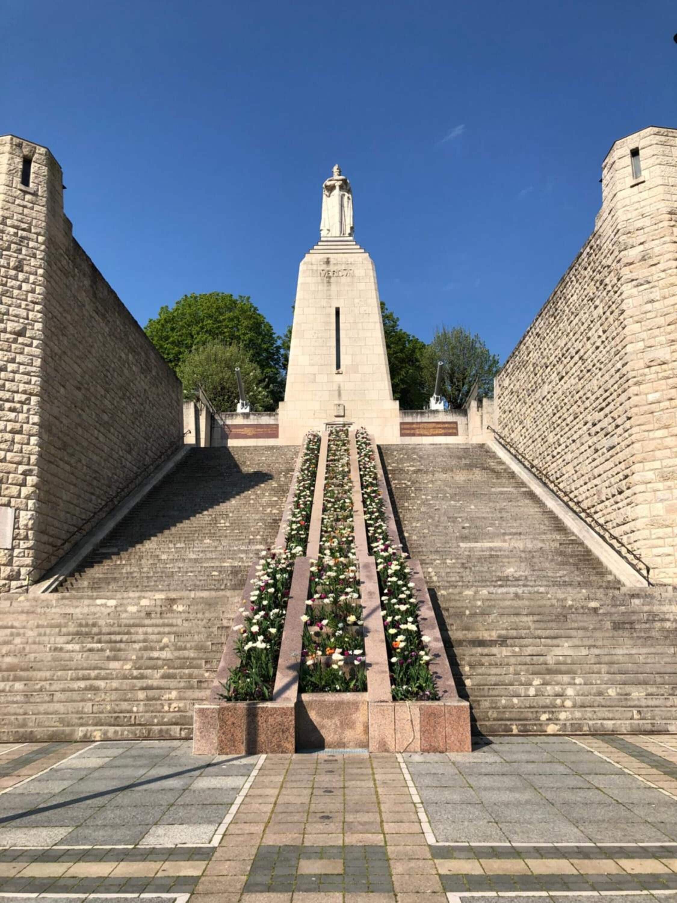 A stone monument with a statue at the top, surrounded by a garden with flowers and a paved pathway leading up to it.