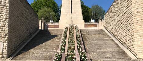 Un monumento de piedra con una estatua en la parte superior, rodeado por un jardín con flores y un camino pavimentado que conduce hasta él.