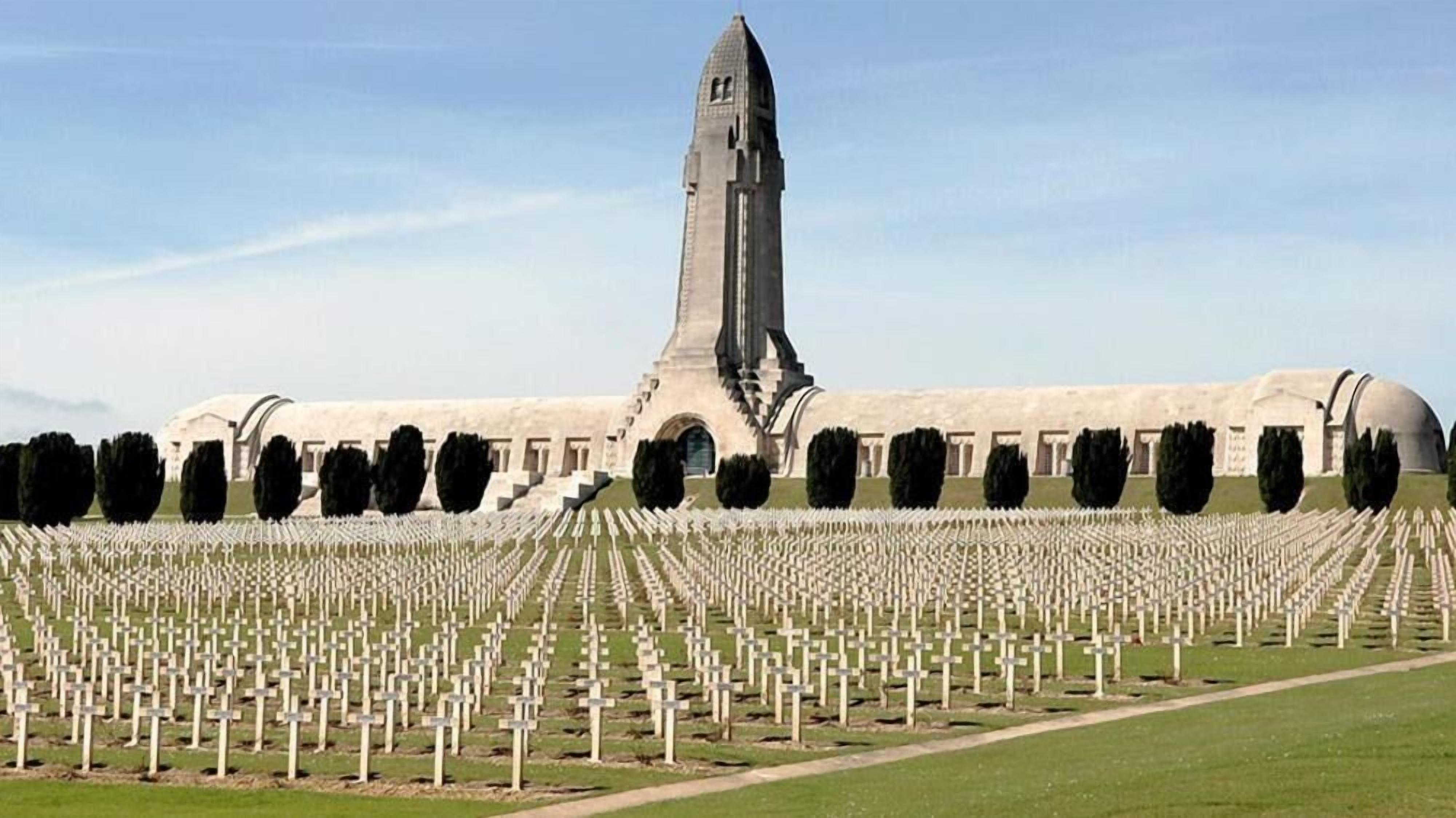 A cemetery with numerous graves arranged in rows, a prominent monument in the center, and a large building in the background.