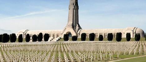 A cemetery with numerous graves arranged in rows, a prominent monument in the center, and a large building in the background.