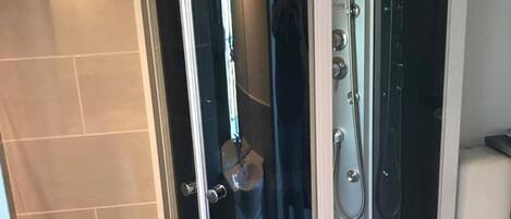 Un receveur de douche d’angle avec une porte en verre, une douchette à main et un panneau de commande fixé au mur.