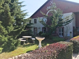 Um hotel com fachada vermelha e branca, área de estar externa e um jardim com diversas plantas e um elemento decorativo em pedra.