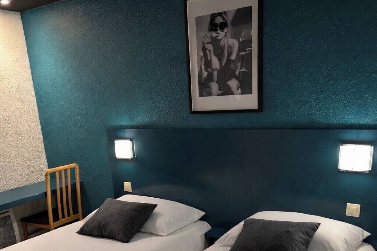 Habitación de hotel con dos camas, cada una con ropa blanca y almohadas grises oscuras. Encima de las camas hay un cuadro enmarcado en la pared.