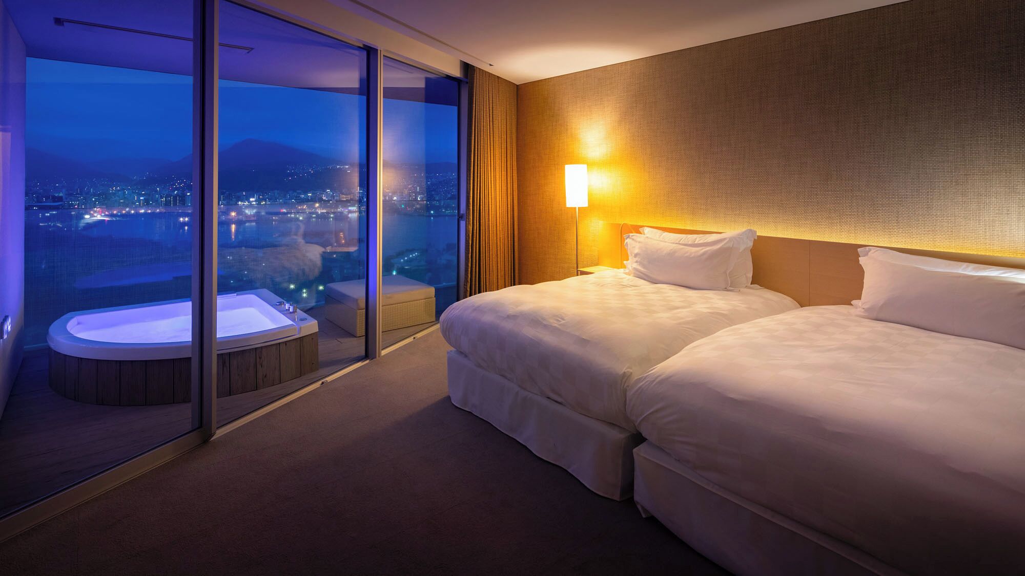 A hotel room with a large sliding glass door, a hot tub, two beds, and a city view.