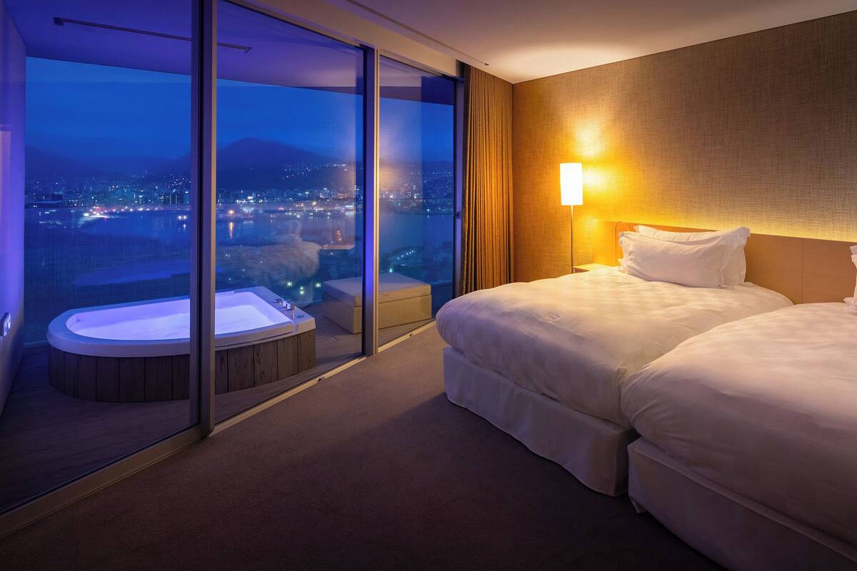 A hotel room with a large sliding glass door, a hot tub, two beds, and a city view.