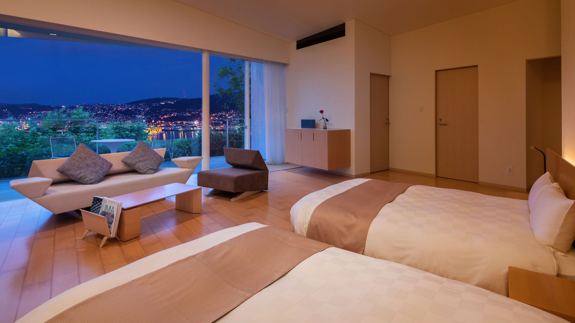 A modern bedroom with a large bed, a sofa, and a chair. There is a view of a cityscape through large windows.