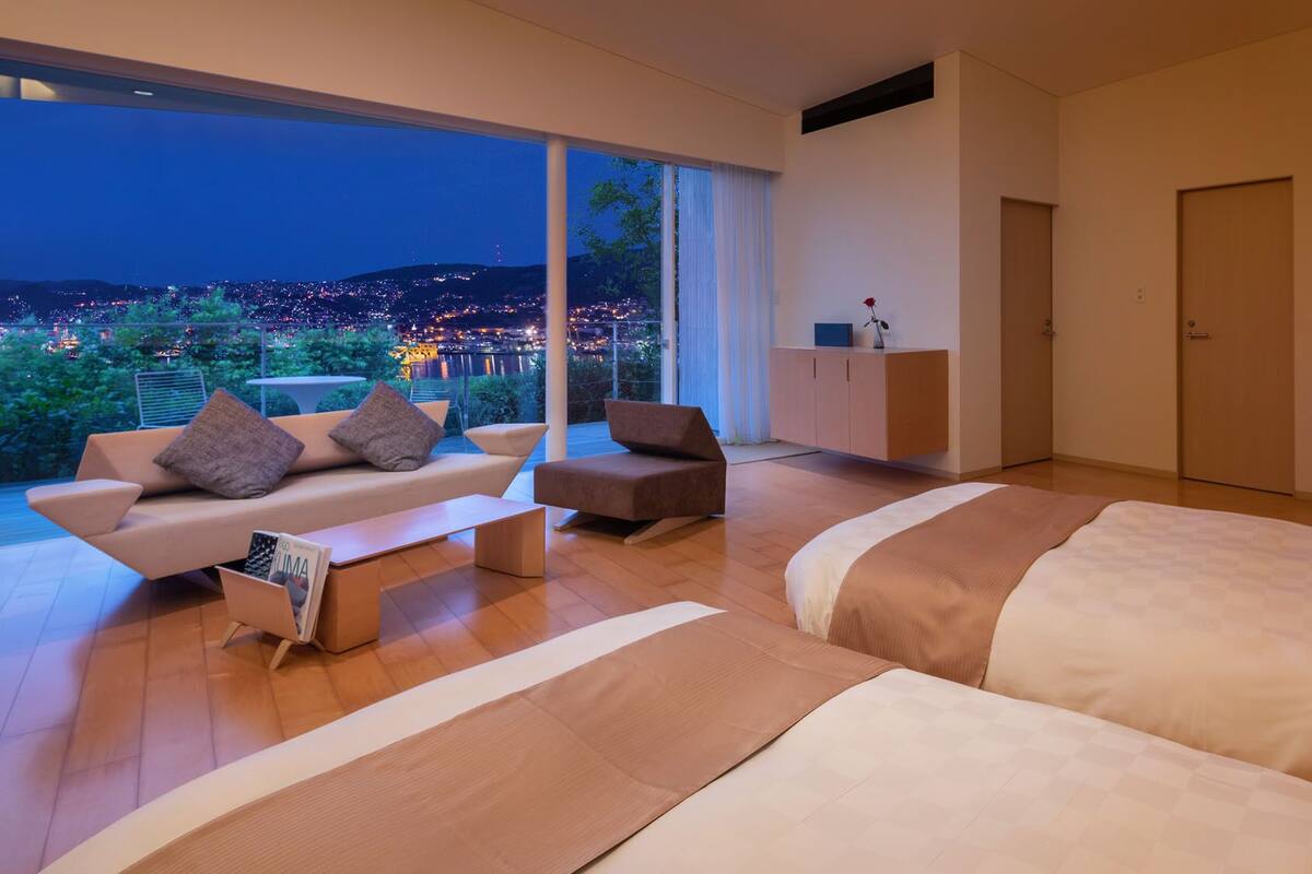 A modern bedroom with a large bed, a sofa, and a chair. There is a view of a cityscape through large windows.
