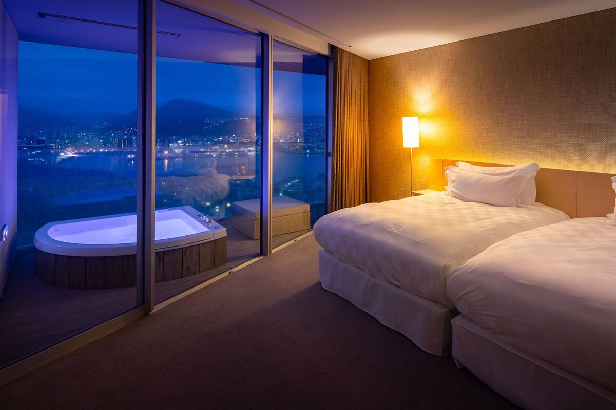 A hotel room with a large sliding glass door leading to a balcony with a hot tub, two beds, and a city view at night.