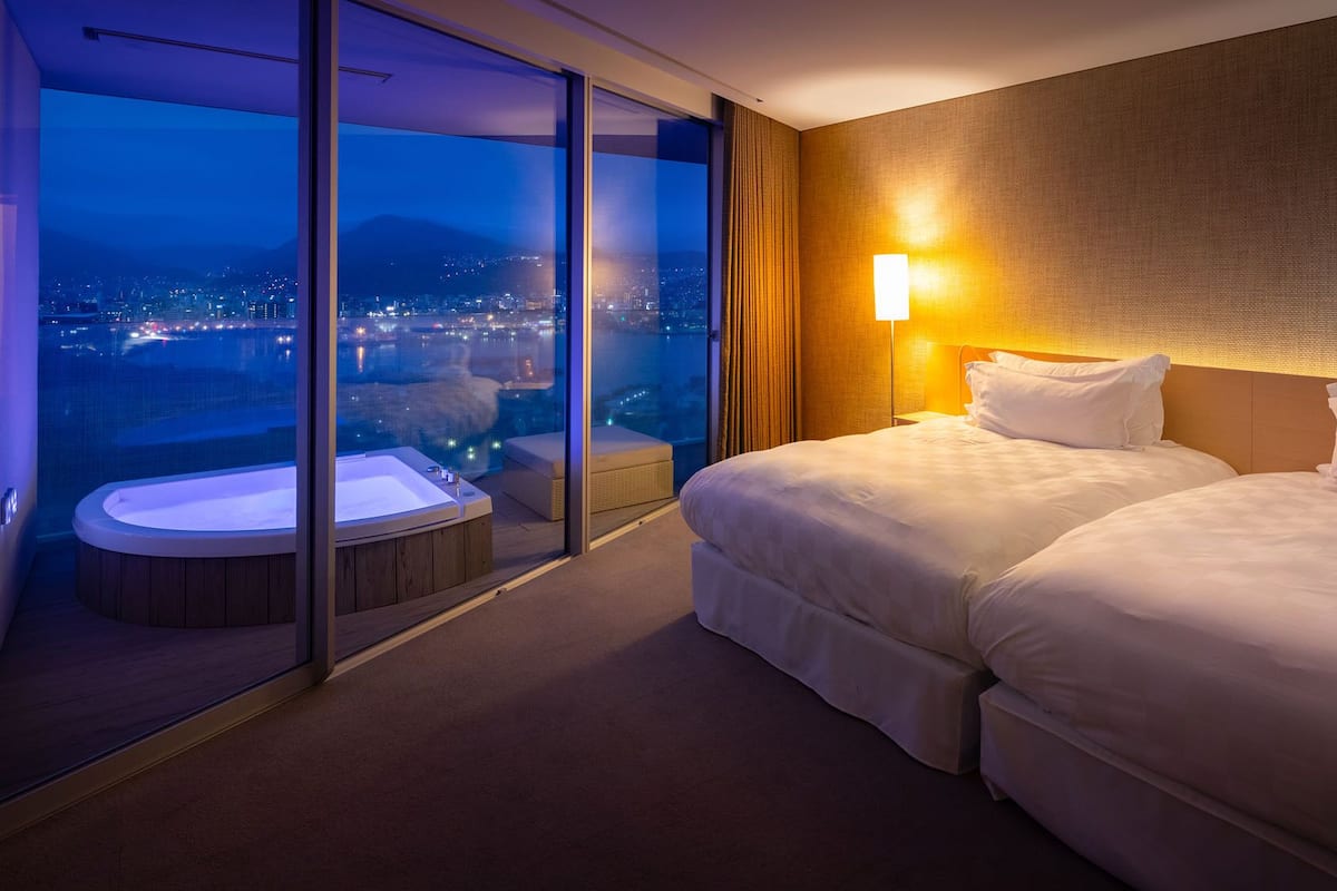 A hotel room with a large sliding glass door leading to a balcony with a hot tub, two beds, and a city view at night.