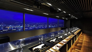 Un long bar avec de nombreux verres et assiettes, une vue sur la ville à travers de grandes fenêtres et un décor intérieur moderne.