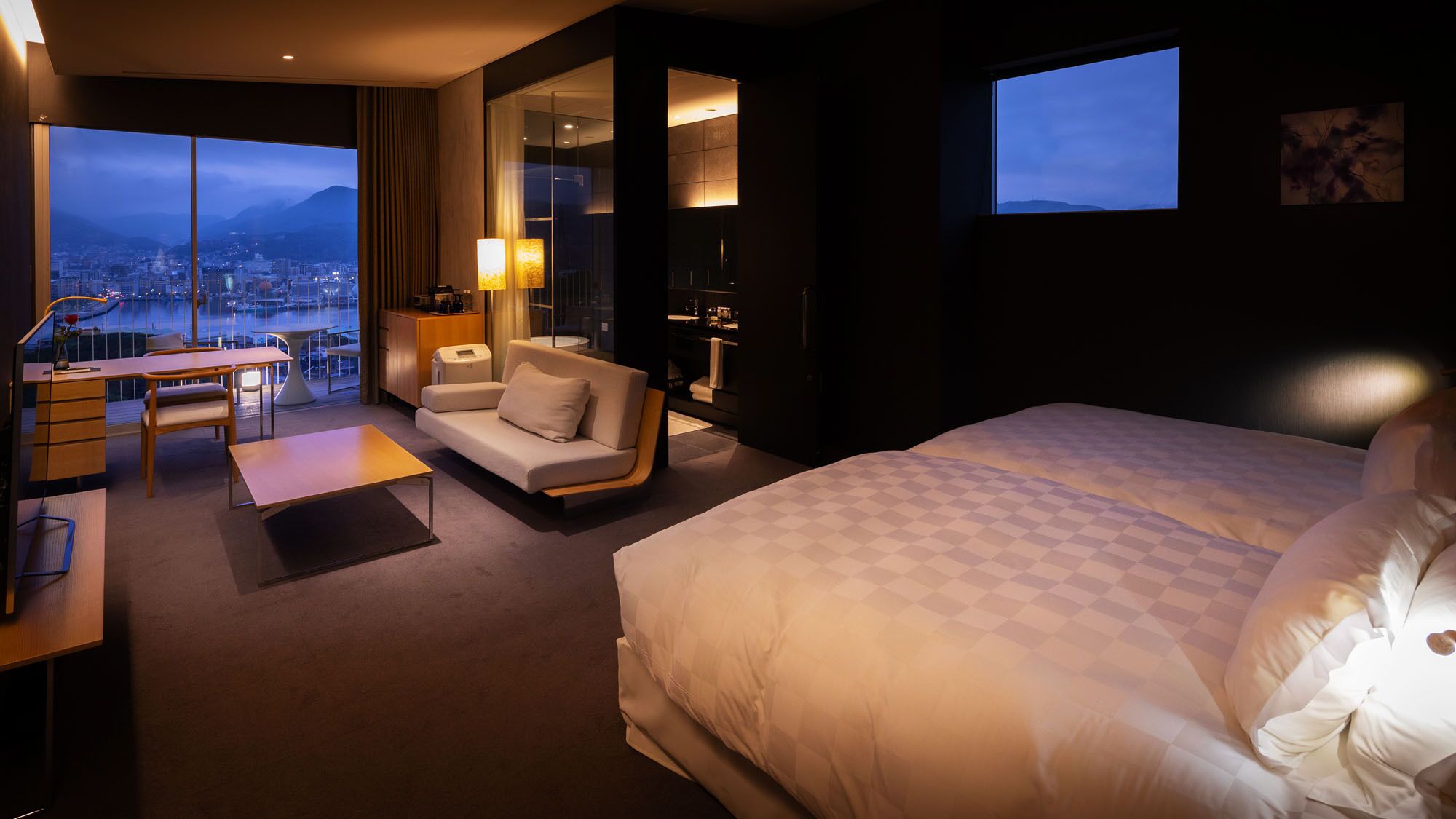 A hotel room with a large bed, a desk with a chair, a sofa, and a view of a cityscape through large windows.