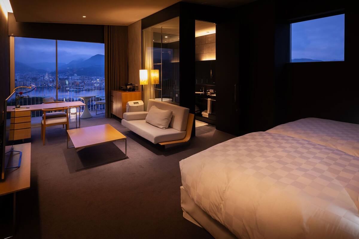 A hotel room with a large bed, a desk with a chair, a sofa, and a view of a cityscape through large windows.
